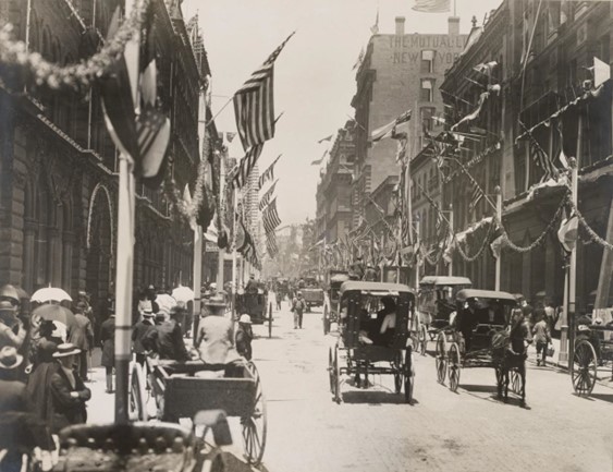A black and white image of a street at the turn of the century. There are people walking along the footpath with horse-drawn carts going up and down the street.