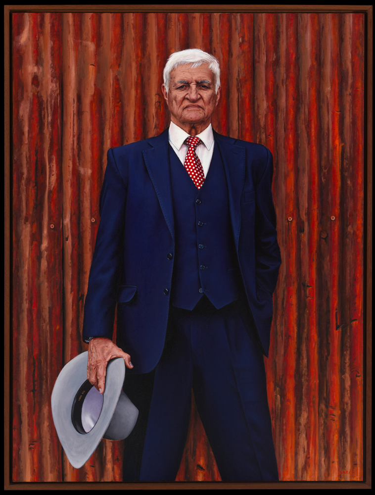 Portrait of The Hon. Robert (Bob) Carl Katter standing in front of a corrugated metal backdrop, wearing a navy blue suit, red polka-dotted tie, and holding a light grey Akubra.